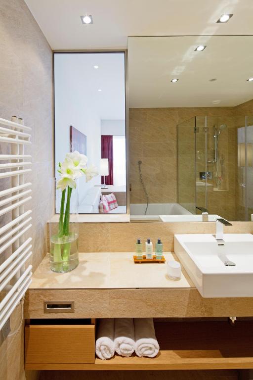 Mūsdienīgs vannas istabas interjers ar elegantu izskatu, kurā ir ziedu kompozīcija, higiēnas līdzekļi un dvieļi. Gaismas un spoguļu elementi rada plašuma sajūtu.