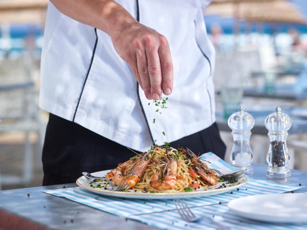 Šefpavārs pievieno garšvielas gardai pasta platei ar garnelēm, kas pasniegta pludmalē. Fons rada relaksētu atmosfēru, piemērotu maltītei brīvā dabā.