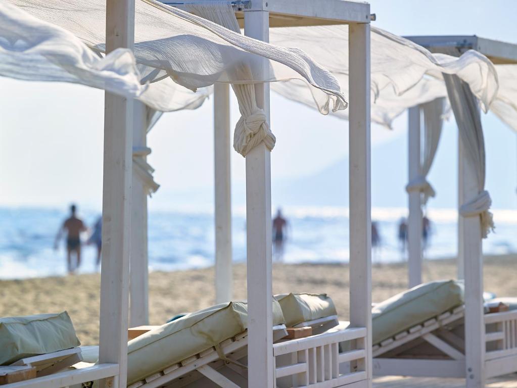 Pludmales kabanas ar baltām drānām un jūras fonu, kurā redzami cilvēki, kas pastaigājas pa pludmali.