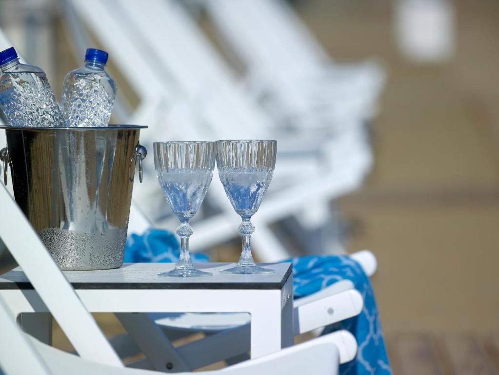 Dzeltenā pludmales krēslā novietots galdiņš ar divām elegantiem glāzēm un ledus spaini, kurā atrodas ūdens pudeles, radot relaksējošu atmosfēru pludmales vidē.