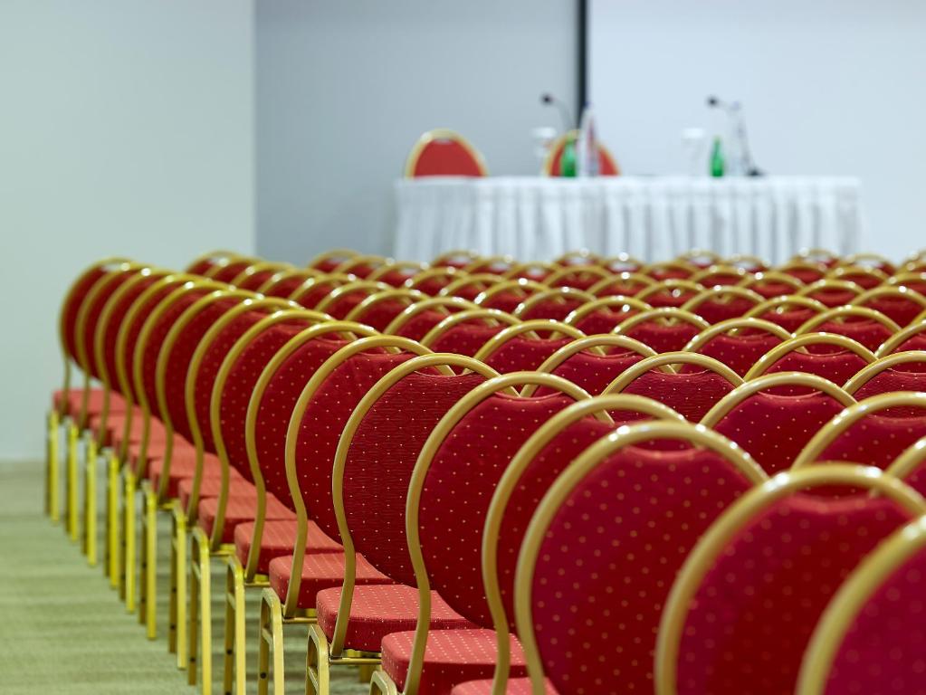 Rindas ar sarkaniem krēsliem konferenču zālē, priekšplānā redzama krēslu rinda, bet fona daļā ir galds ar dekoratīviem elementiem.