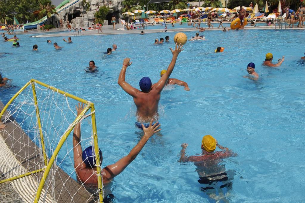 Ūdens polo spēles ainava baseinā, kur spēlētāji aktīvi piedalās spēlē, kamēr citi apmeklētāji izklaidējas. Ūdens ir dzidrs un saulains laiks.