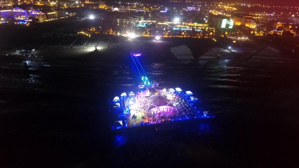 Krāsains nakts skats uz pludmali ar gaismas efektiem un cilvēkiem, kas izklaidējas uz promenādes pie jūras. Vides apgaismojums rada dzīvīgu atmosfēru.