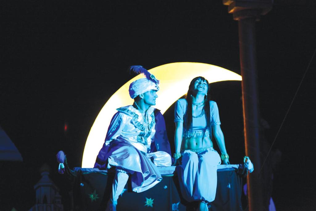 Izpildītāji, tērpušies krāsainos kostīmos, sēž uz skatuves zem liela, spoža mēness. Atmosfēra ir maģiska un noslēpumaina, radot sajūtu par pasakainu stāstu.