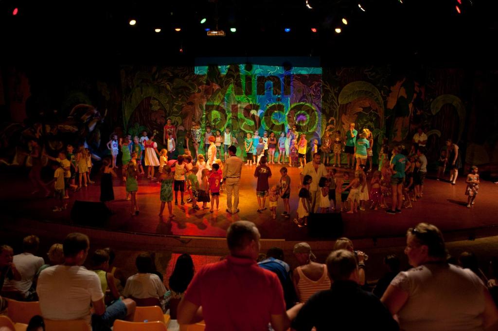 Bērnu diskotēka ar daudziem bērniem, kas dejo uz skatuves, apkārt pieaugušie skatītāji. Aiz skatuves ir krāsaini zīmējumi, kas veido jautru atmosfēru.