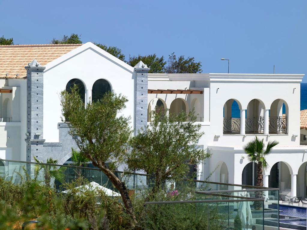 Balta, moderna villa ar arhitektūras elementiem, piemēram, arkas un kolonām, ieskauta zaļumiem un palmu kokiem, ar skatu uz jūru.