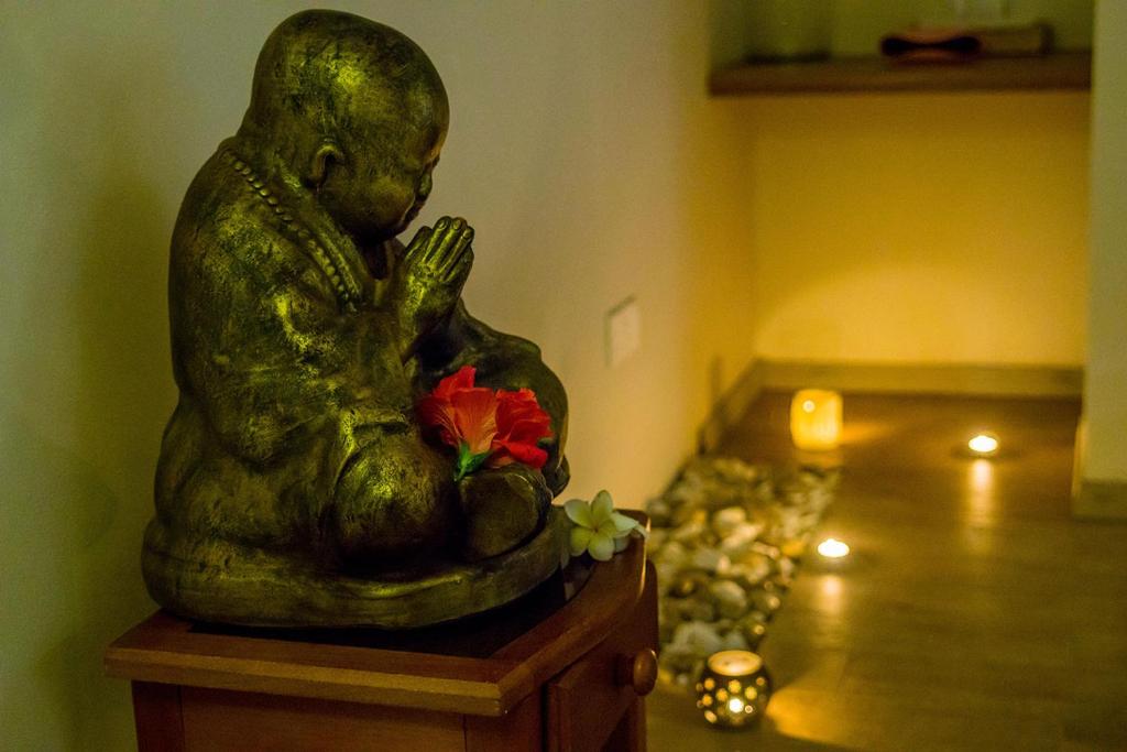Zelta statuja, kas attēlo meditējošu budu, ir novietota uz koka skapīša, blakus ziediem un svecēm, radot mierīgu atmosfēru.
