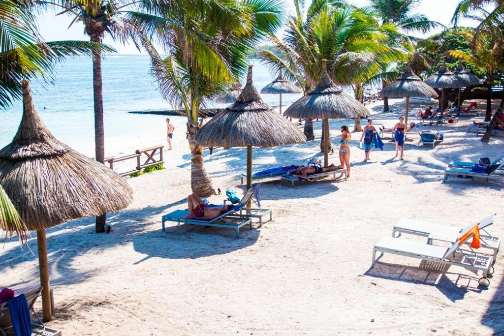 Pludmales ainava ar palmām un sauļošanās krēsliem, kur cilvēki atpūšas un izbauda sauli. Ūdens ir skaidrs un mierīgs, radot relaksējošu atmosfēru.