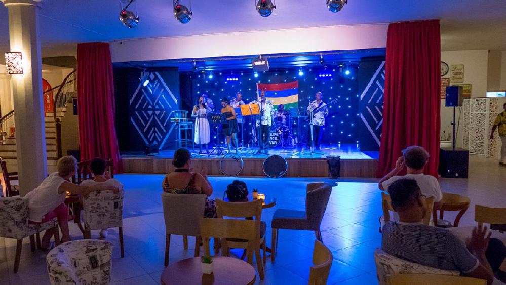 Dzīvās mūzikas pasākums ar grupu, kas uzstājas uz skatuves, apgaismota ar zilu gaismu. Apmeklētāji sēž pie galdiņiem, baudot izrādi un atmosfēru.