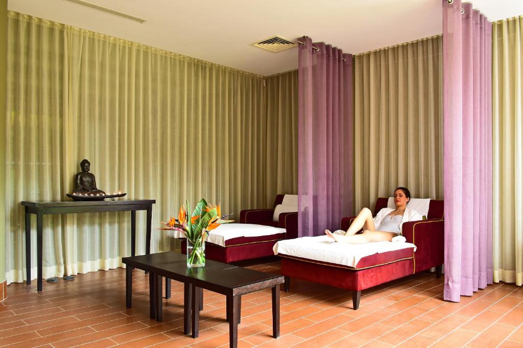 Relaksējoša spa telpa ar diviem sarkaniem atpūtas krēsliem un ziediem uz galda, kur sieviete atpūšas. Telpu rotā dzeltenas un purpursarkanas aizskari, kā arī Budas statuja.