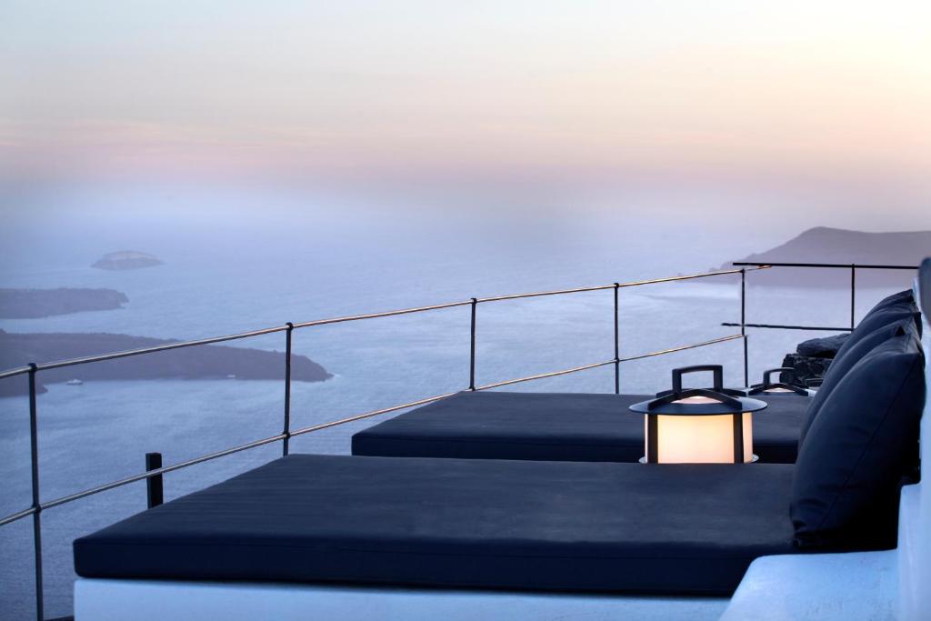 Ainava ar skatu uz jūru, kur divas atpūtas gultas ir izvietotas uz terases, ar laternu starp tām. Gaisma un krāsas rada mierīgu atmosfēru saulrieta laikā.