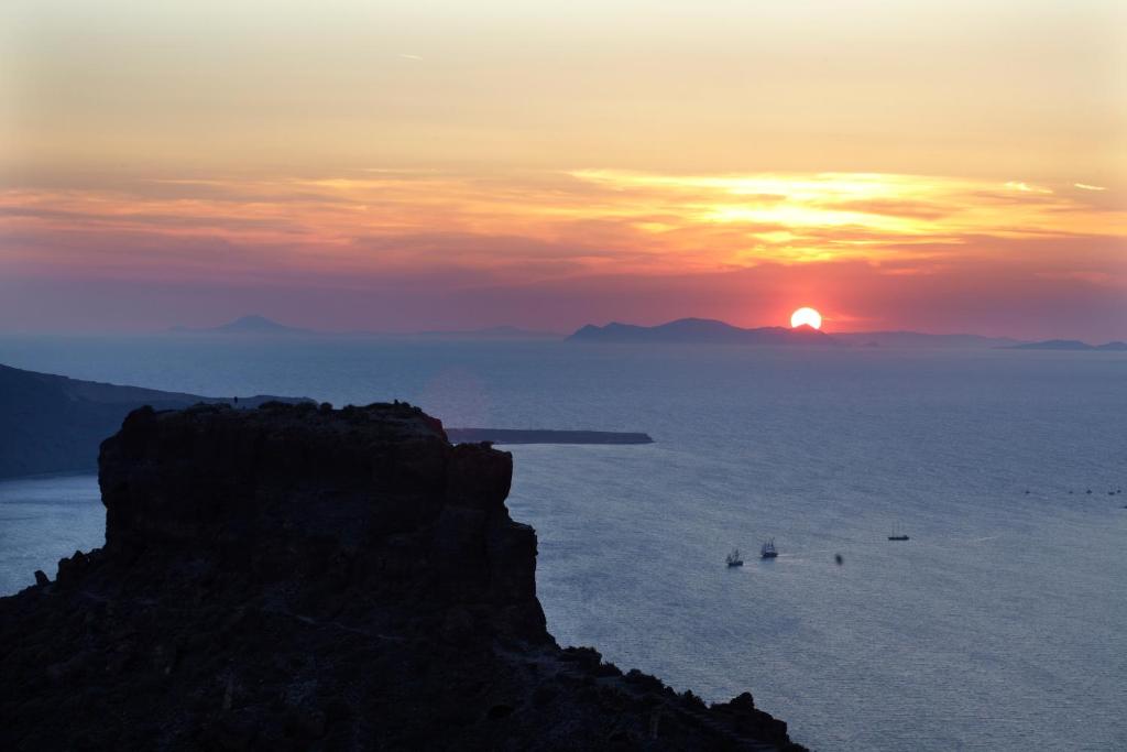 Saule rietot pāri jūrai, radot krāsainu debesu ainavu un klints siluetu priekšplānā.