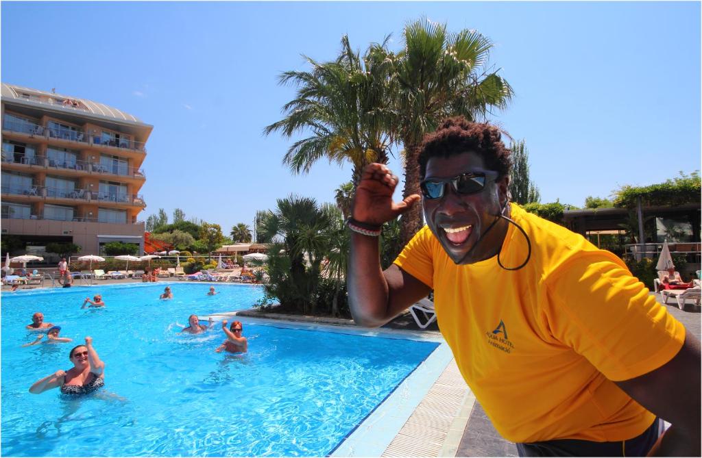 Priecīgs darbinieks pie baseina, sveicina viesus siltā saulainā dienā. Fons ietver viesnīcu un peldētājus, kas izbauda ūdeni.