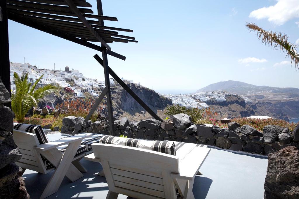 Skats uz Santorini no terases ar baltām koka mēbelēm un zaļiem augiem, fons ar kalnu un jūras ainavu.
