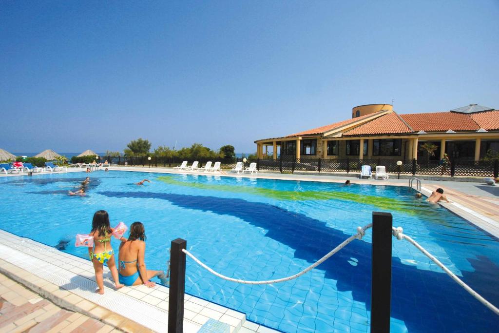 Baseins ar bērniem un pieaugušajiem, kas izbauda saulainu dienu. Apkārt baseinam redzami sauļošanās krēsli un skaista ainava.