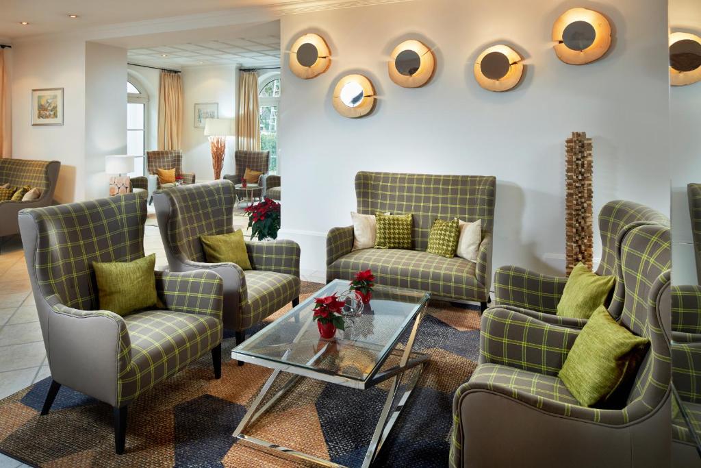 Mūsdienīgs viesnīcas interjers ar ērtiem krēsliem un dīvānu, rotātiem ar spilveniem, un stikla galdu. Sienas rotā dekoratīvi elementi un gaismas.