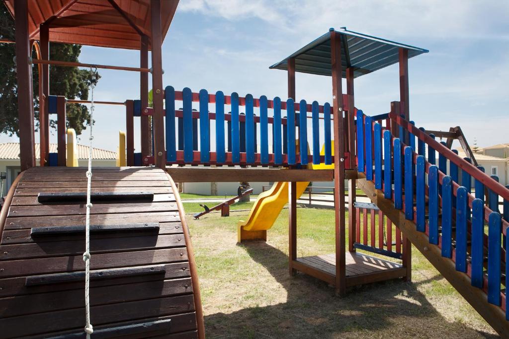 Bērnu spēļu laukums ar koka struktūru, slīdkalniņu un kāpnēm, kas aicina bērnus rotaļāties un izpētīt. Spēļu laukuma krāsainās sētas un jumts piešķir dzīvīgu atmosfēru.