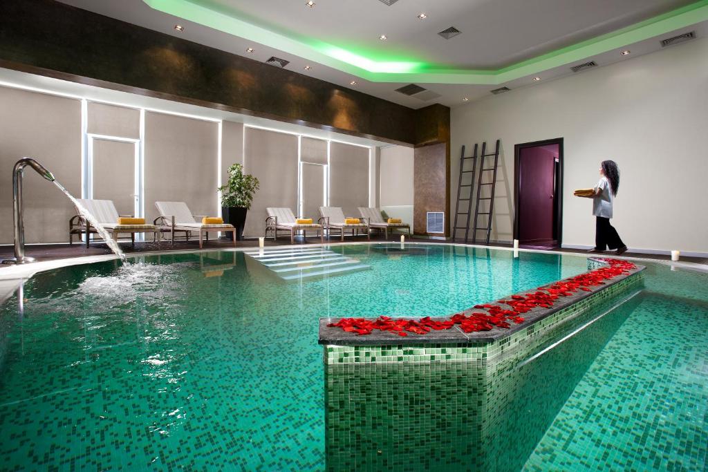 Mūsdienīga spa telpa ar zaļu apgaismojumu, baseinu un atpūtas krēsliem. Ūdens plūst no izlietnes, un uz baseina malas ir ziedlapiņas.