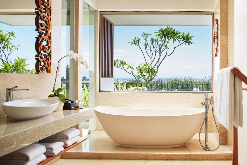 Mūsdienīgs vannas istabas interjers ar elegantu brīvi stāvošu vannu un skatu uz dabu caur lielu logu. Dabas elementi un gaismas krāsas rada relaksējošu atmosfēru.