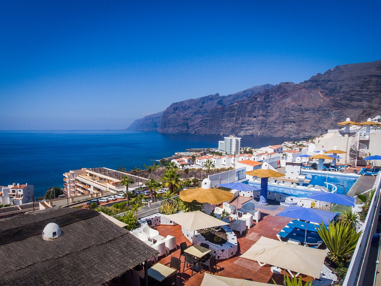 Viesnīcas terase ar skatu uz okeānu un kalniem, kurā redzami baseini un atpūtas zonas. Skaists laiks un zila debesis papildina šo idillisko ainavu. panoramic view with beach and pools