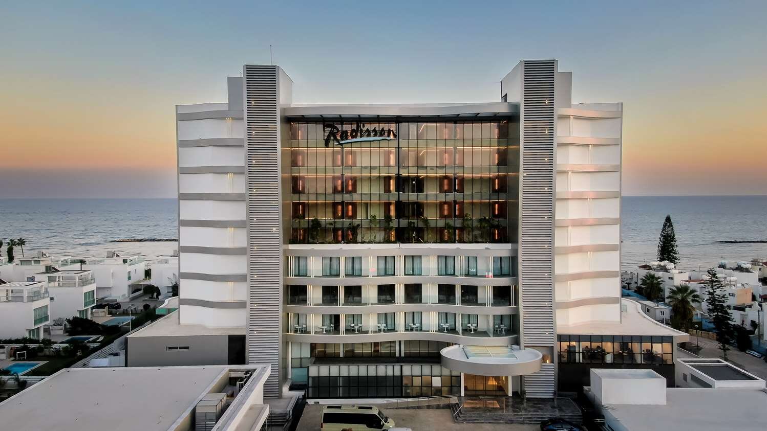 Mūsdienīgs Radisson viesnīcas eksterjers ar skatu uz jūru, kas atspoguļo saulrietu.