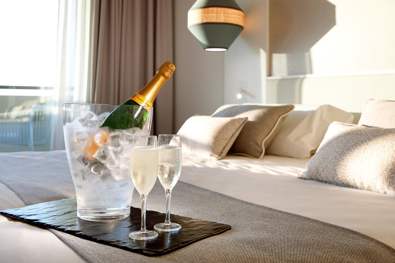 Šampanieša pudele ledus traukā kopā ar divām šampanieša glāzēm uz gultas, kas rotāta ar mīkstām spilveniem un modernu apgaismojumu.