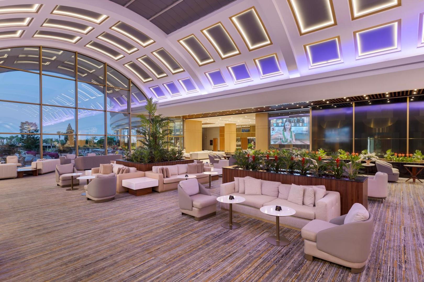 Miracle Resort Hotel lobby bārs ar mīkstajiem dīvāniem, dekoratīviem augiem un lieliem logiem, pa kuriem paveras skats uz dārzu vakarā.