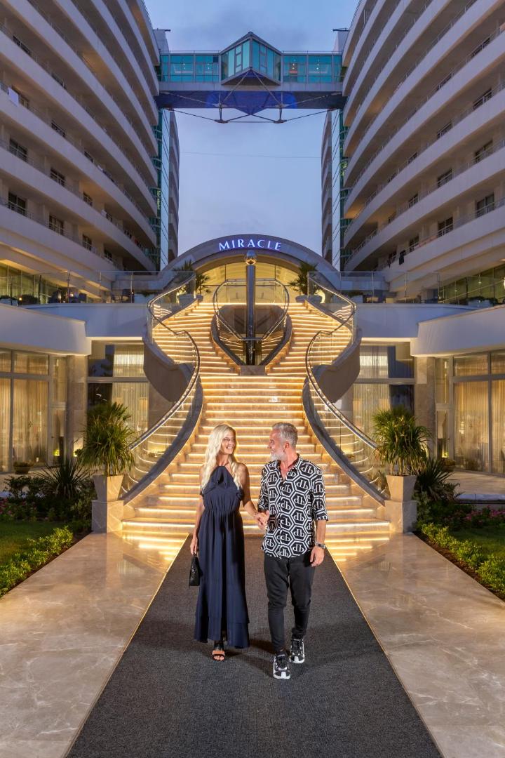 Miracle Resort Hotel izgaismotās centrālās kāpnes vakarā ar pāri priekšplānā, parādot viesnīcas moderno fasādi un eleganto ieeju