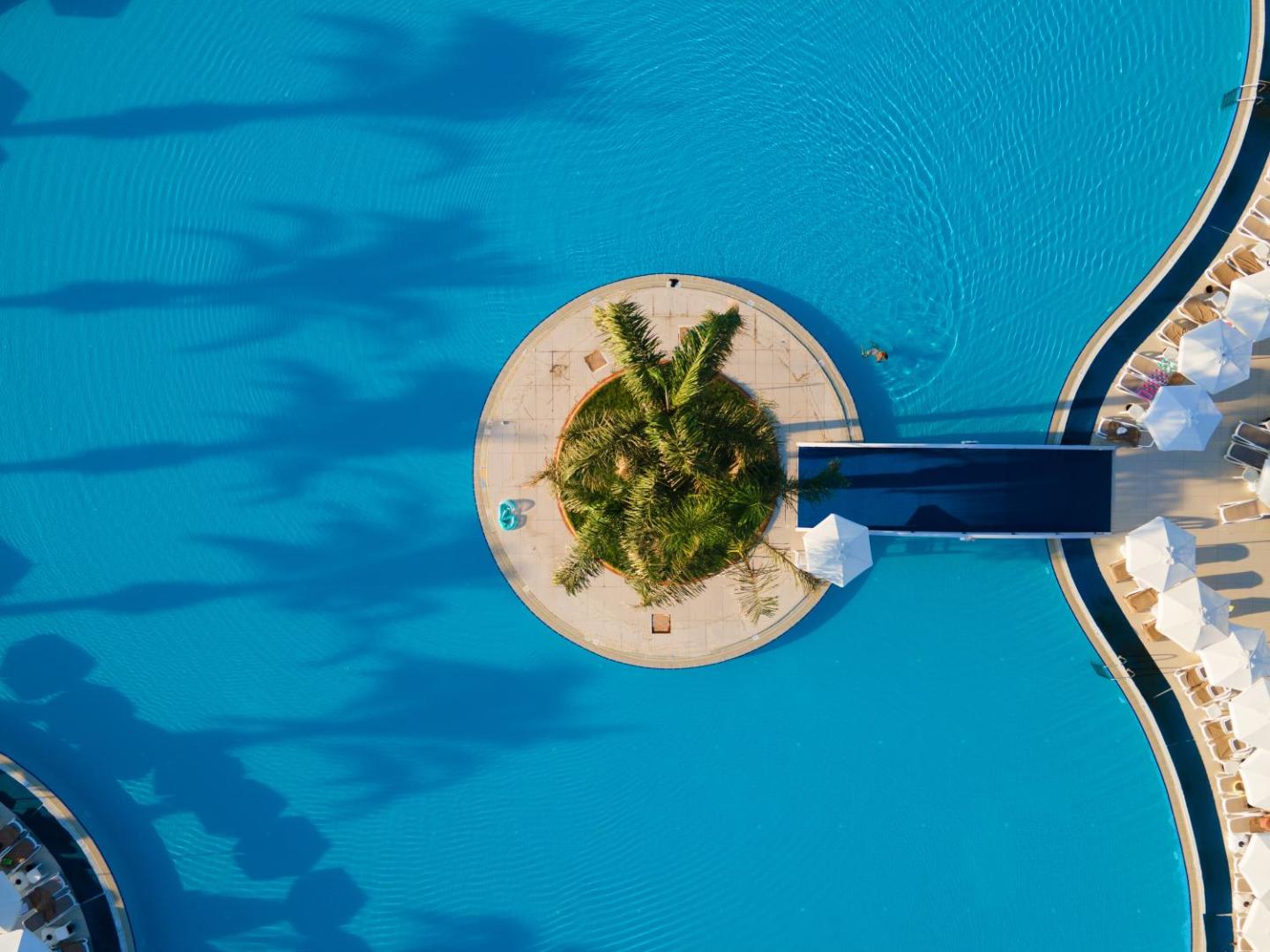 Miracle Resort Hotel baseina detaļa no augšas — maza palmu sala baseina vidū ar tiltiņu un tirkīzzilu ūdeni
