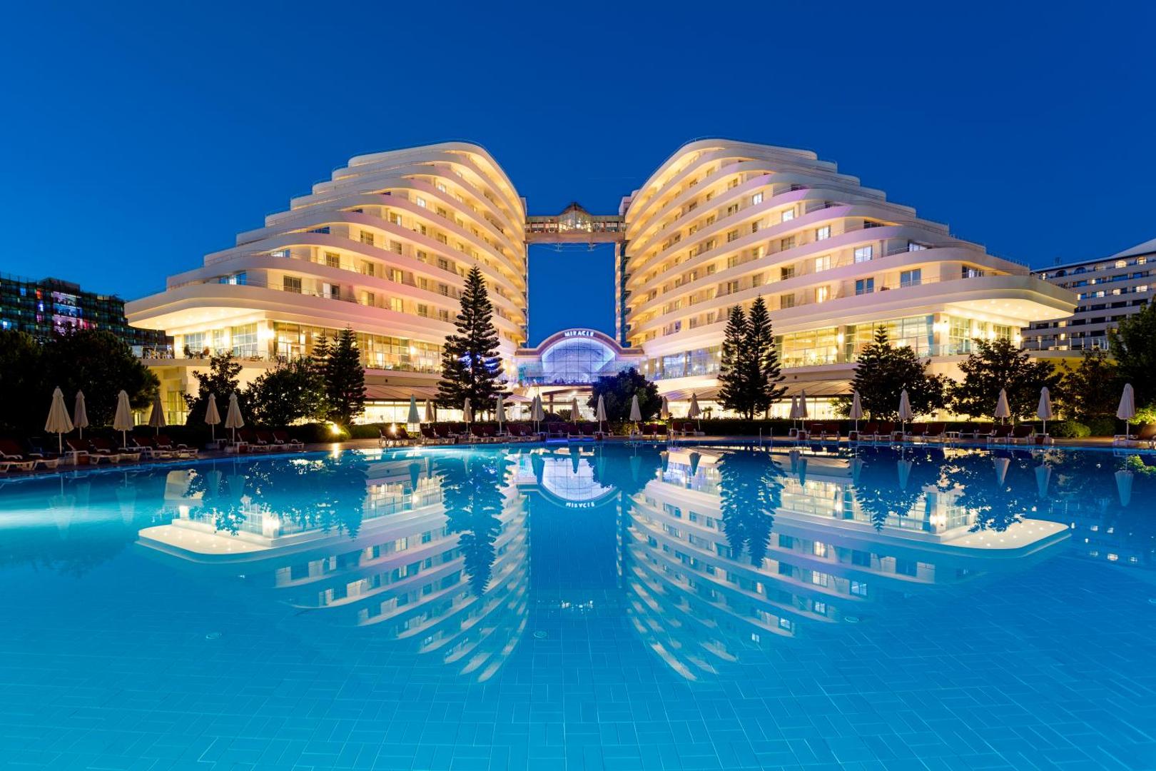 Miracle Resort Hotel izgaismotā fasāde un baseins vakarā Antalijā — spoguļattēls ūdenī un mājīgs apgaismojums uz balkoniem