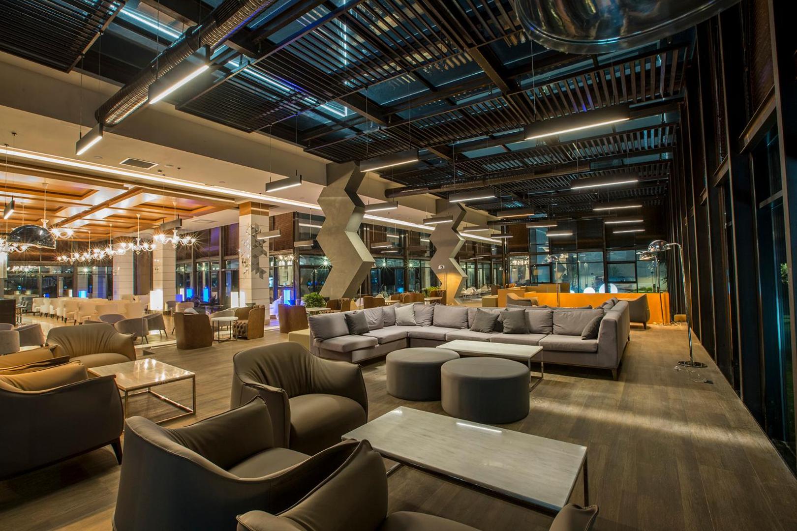 Numa Bay Exclusive Hotel modernā lobby atpūtas zona vakarā ar mīkstajiem dīvāniem, krēsliem un industriāla stila griestu dizainu Alanjā