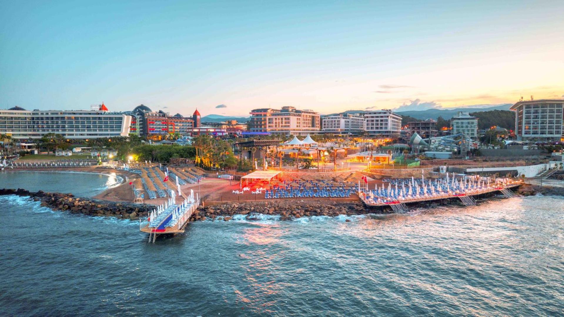 Numa Bay Exclusive Hotel panorāmas skats uz piekrasti saulrietā — apgaismota pludmales zona, piestātne un viesnīcas korpusi Alanjas reģionā