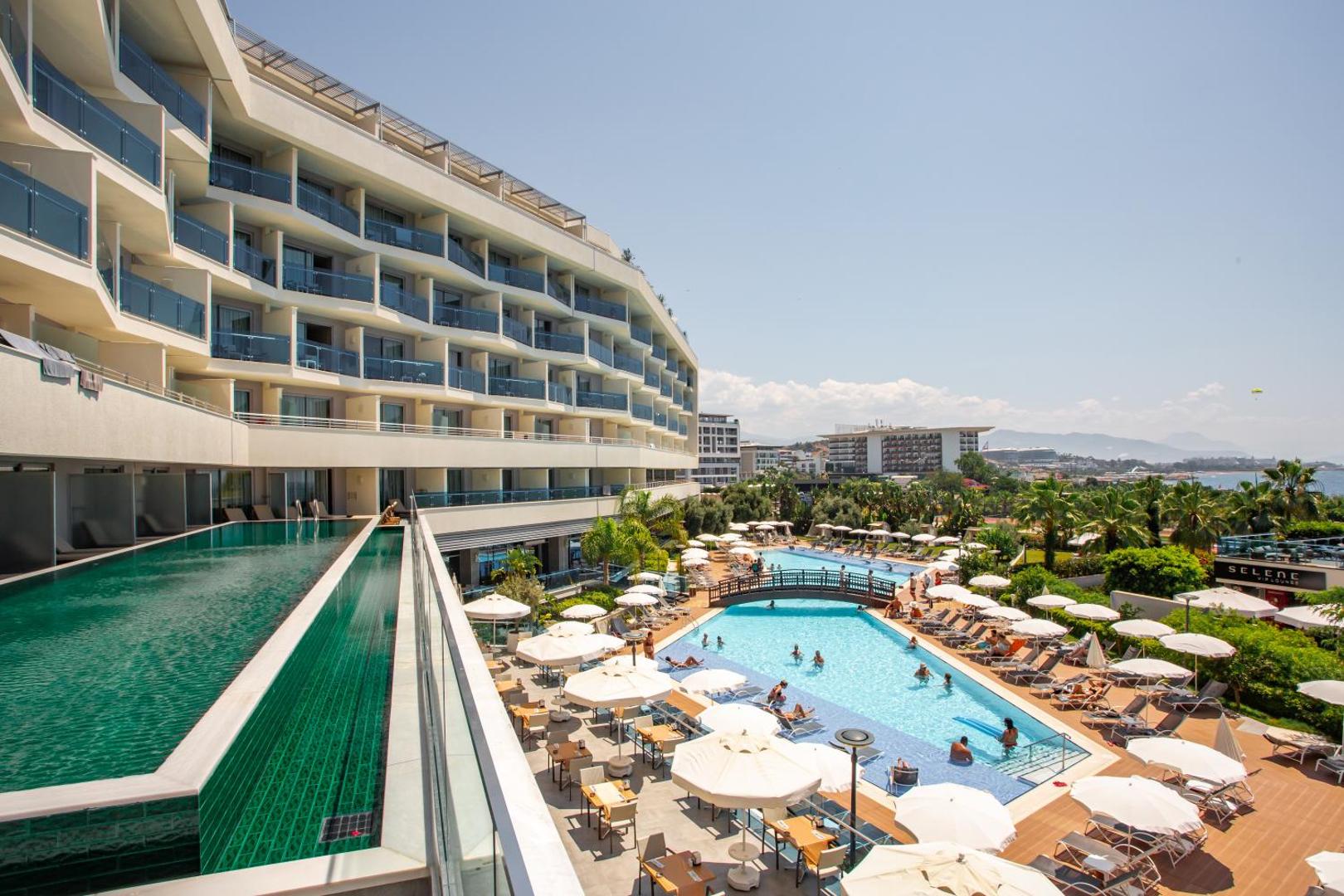 Selene Beach and Spa Hotel ēkas fasāde un lielais āra baseins ar saulessargiem, tiltiņu un palmu dārzu Alānijas piekrastē