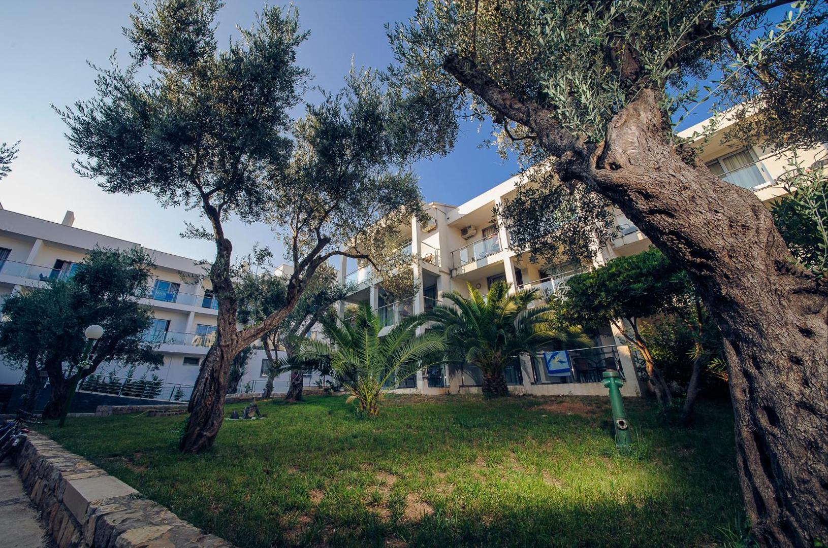 Viesnīcas Vile Oliva numuru korpuss ar balkoniem, ko ieskauj zaļš zāliens un veci olīvkoki saulainā dienā