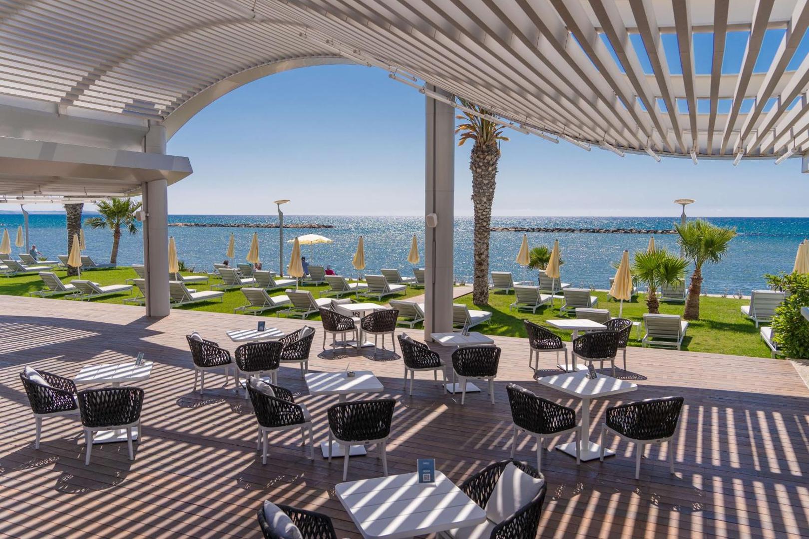 Radisson Beach Resort Larnaca āra terase ar moderniem krēsliem un galdiņiem zem nojumes, skats uz zālienu un jūru
