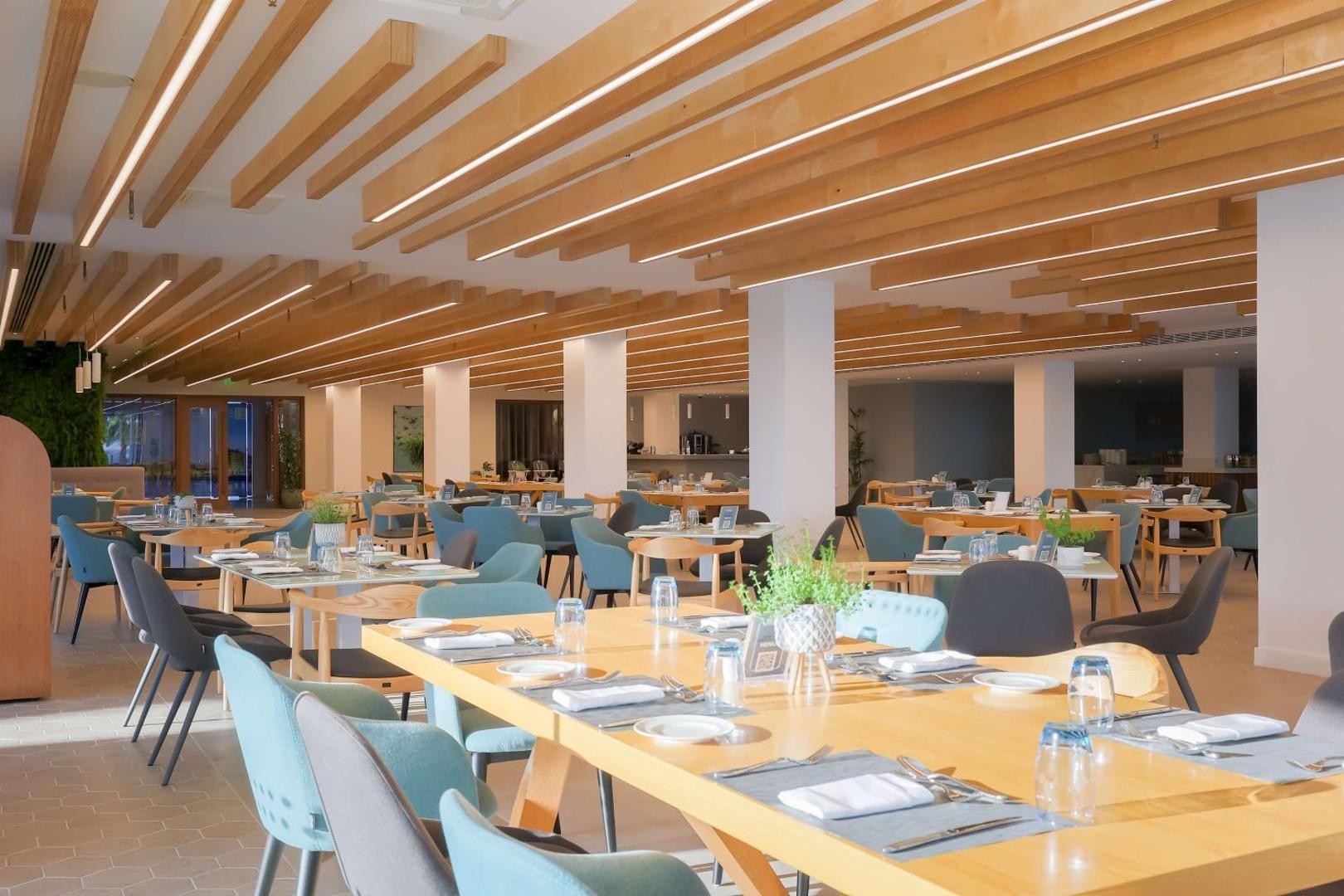 Radisson Beach Resort Larnaca plaša brokastu zāle ar gaiša koka griestu sijām, ziliem krēsliem un servētiem galdiem