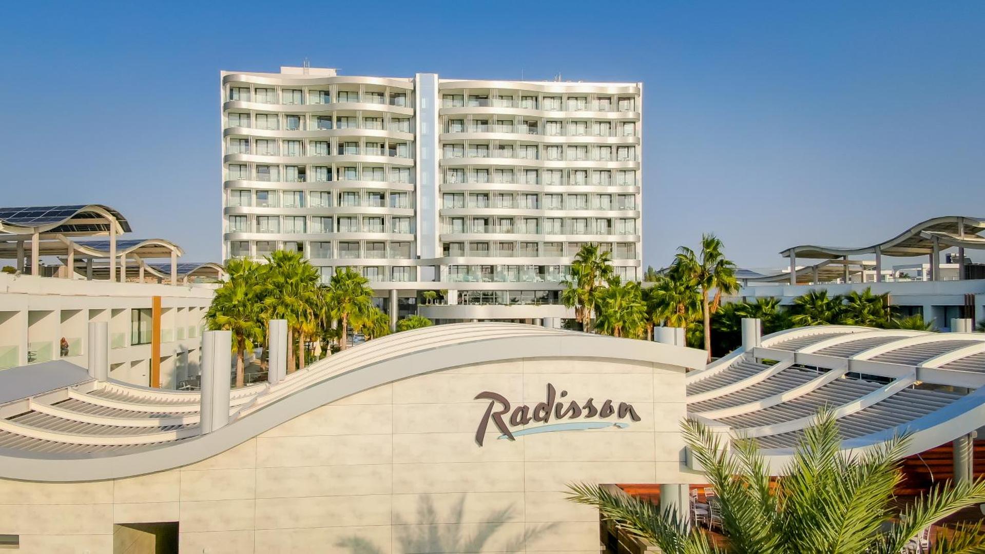 Radisson Beach Resort Larnaca galvenā ēka un zemākie numuru korpusi dienas gaismā — gaiša arhitektūra un sakopta teritorija Kiprā