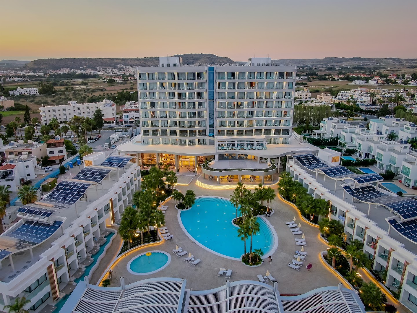 Radisson Beach Resort Larnaca teritorijas skats no augšas — liels āra baseins, bērnu baseins un saules paneļi uz zemāko korpusu jumtiem