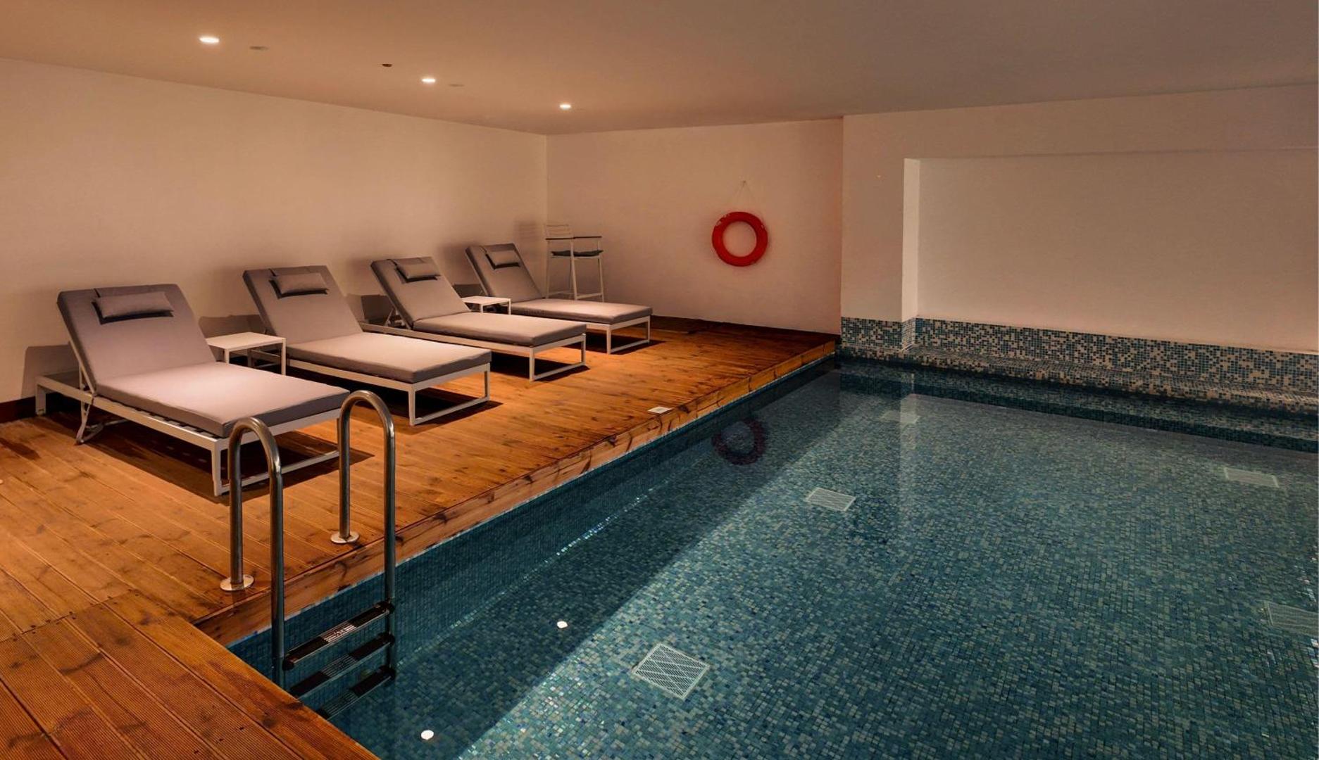 Radisson Beach Resort Larnaca iekštelpu baseins ar koka klāju, pelēkiem sauļošanās krēsliem un mierīgu apgaismojumu spa zonā