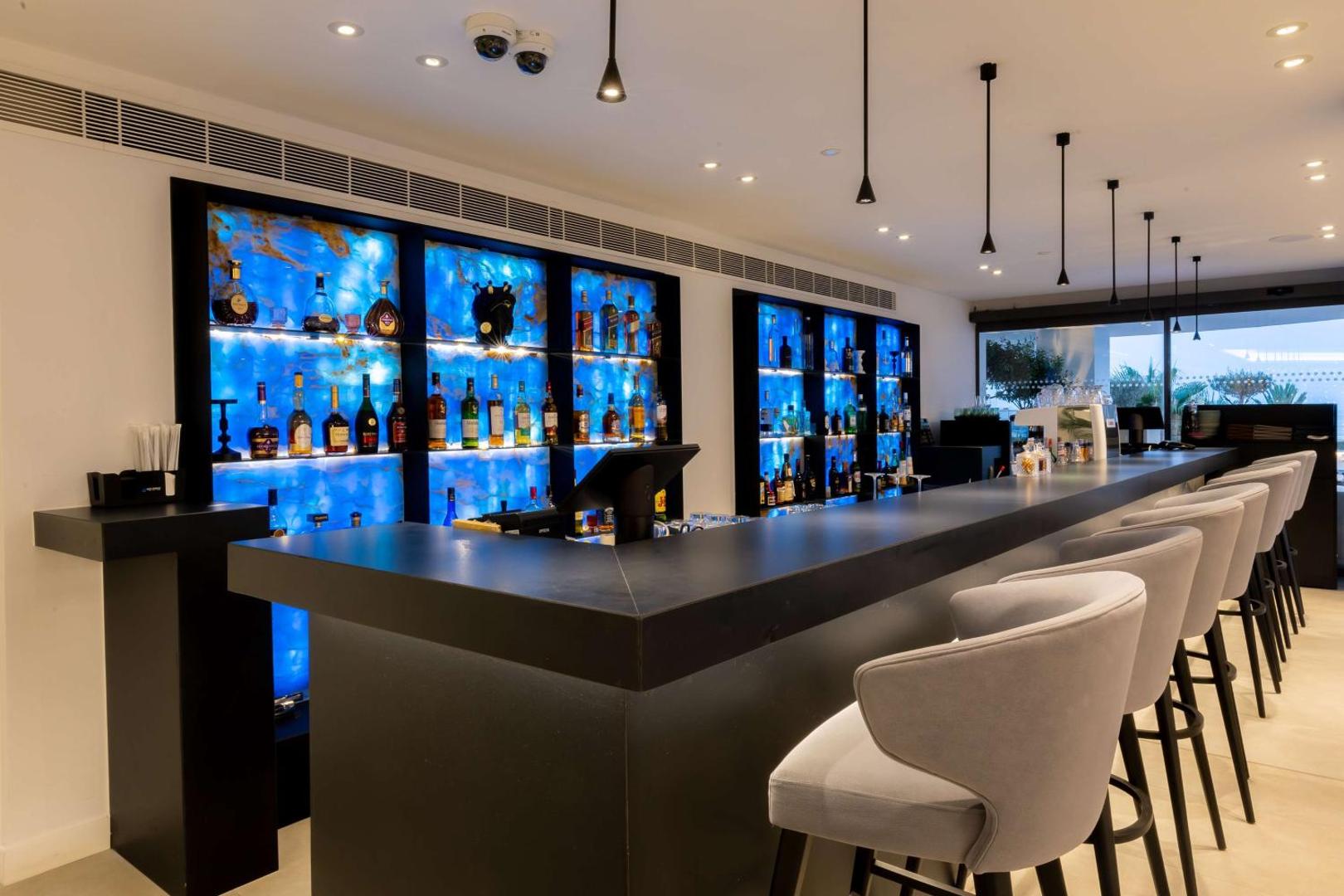 Stilīgs Radisson Beach Resort Larnaca bārs ar izgaismotiem ziliem plauktiem, tumšu leti un elegantiem bāra krēsliem Larnakā.