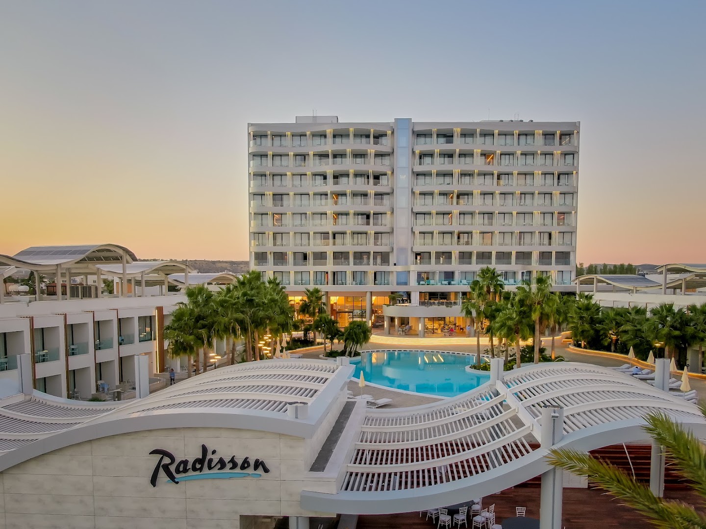 Radisson Beach Resort Larnaca skats uz centrālo baseinu un viesnīcas korpusu vakarā — palmas, atpūtas krēsli un izgaismota terases zona