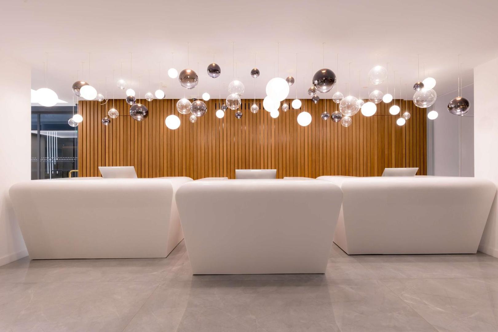 Radisson Beach Resort Larnaca viesnīcas recepcijas zona ar modernām baltām letēm un dekoratīvām stikla lodes lampām pie koka paneļu sienas.