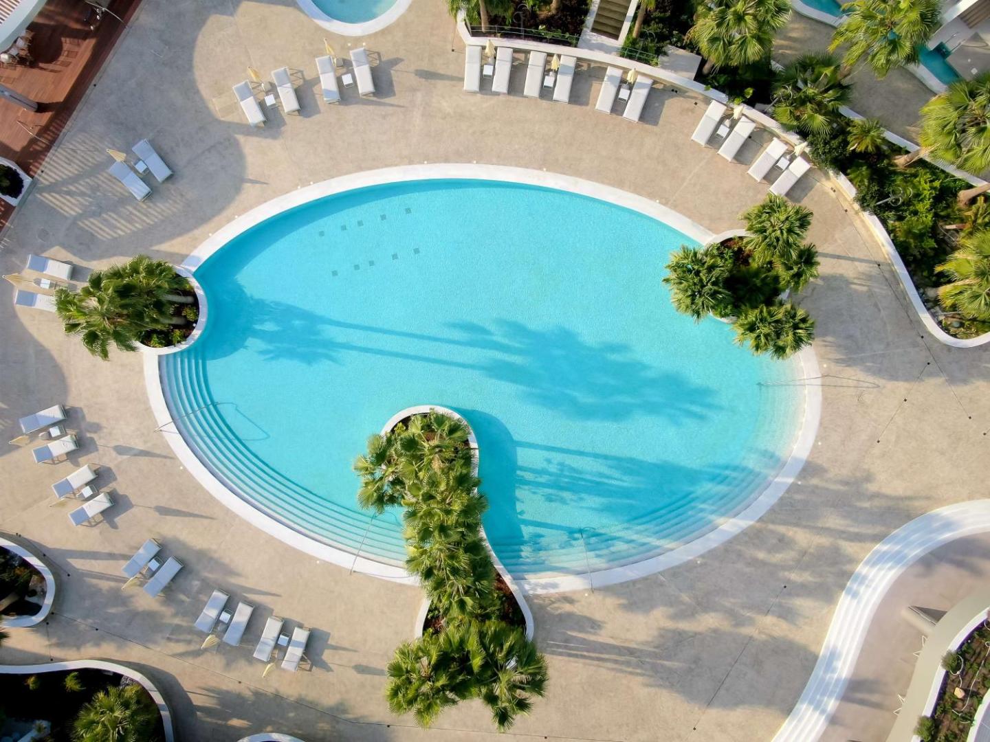 Radisson Beach Resort Larnaca āra baseins no putna lidojuma — ovālas formas baseins ar tirkīza ūdeni, palmām un sauļošanās krēsliem Kiprā