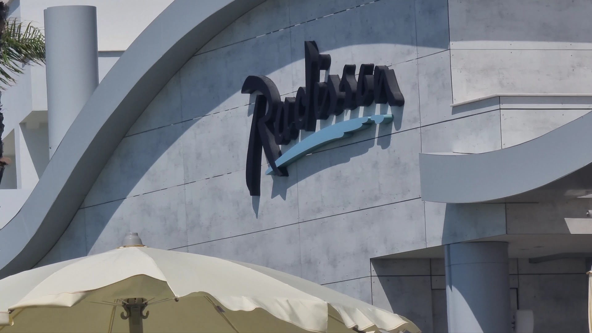 Radisson Beach Resort Larnaca viesnīcas logotips uz modernas fasādes sienas — tuvplāns ar saulessarga malu priekšplānā