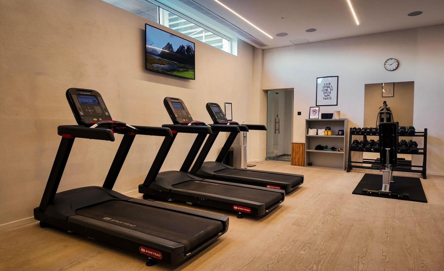 Radisson Beach Resort Larnaca sporta zāle ar skrejceliņiem, hantelēm un TV ekrānu pie sienas — mūsdienīgs fitnesa aprīkojums viesiem
