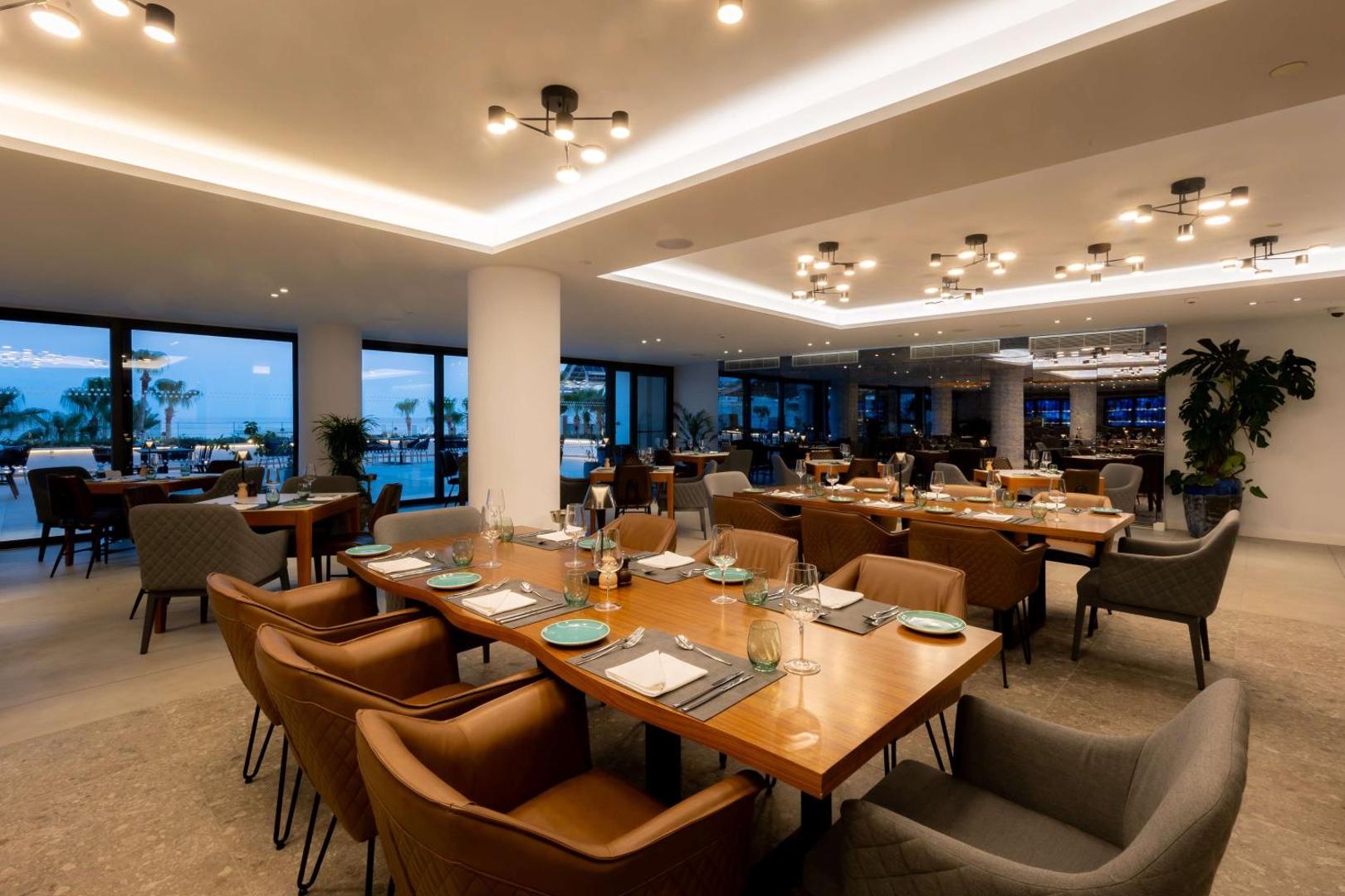 Radisson Beach Resort Larnaca elegants ēdienkartes restorāns ar modernām mēbelēm, siltu apgaismojumu un skatu uz vakara piekrasti Larnakā