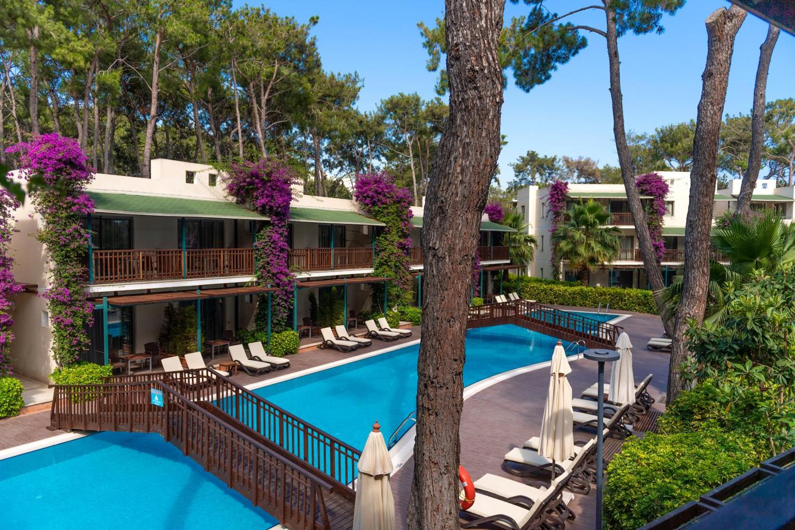 Turquoise Hotel bungalo tipa numuri pie baseina priežu meža ielokā Sidē — terases ar ziedošiem augiem un koka tiltiņš pār baseinu