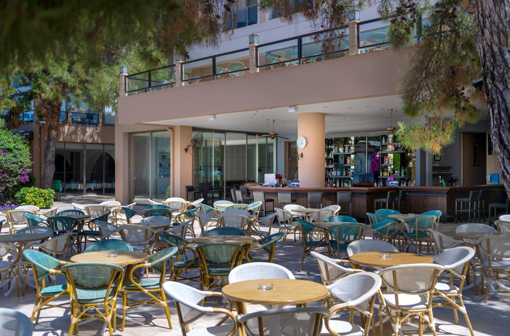 Turquoise Hotel āra terases bārs ar daudziem galdiņiem un krēsliem priežu koku pavēnī blakus viesnīcas galvenajai ēkai