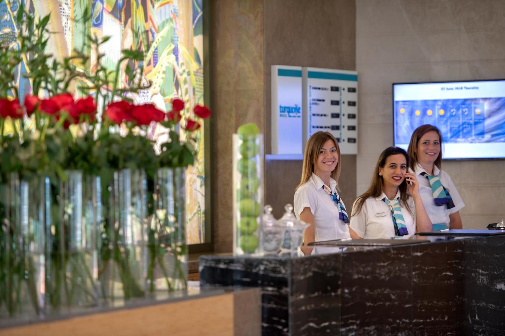 Turquoise Hotel reģistratūras darbinieces pie letes modernā viesnīcas vestibilā ar ziedu dekorācijām fonā.