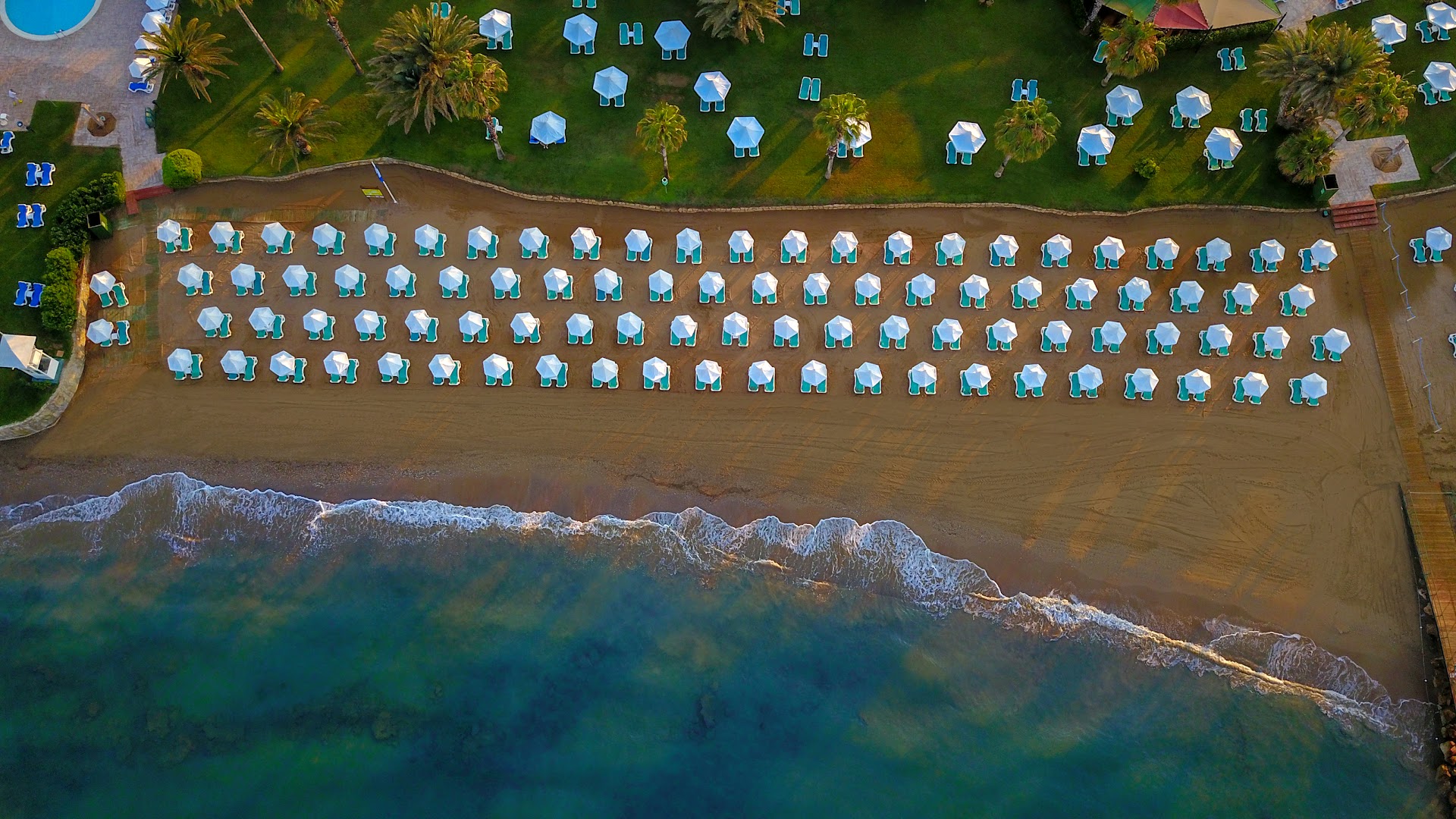 Turquoise Hotel pludmales josla no augšas ar simtiem sakārtotu saulessargu un krēslu pie tirkīzzilā ūdens Sorgunas piekrastē